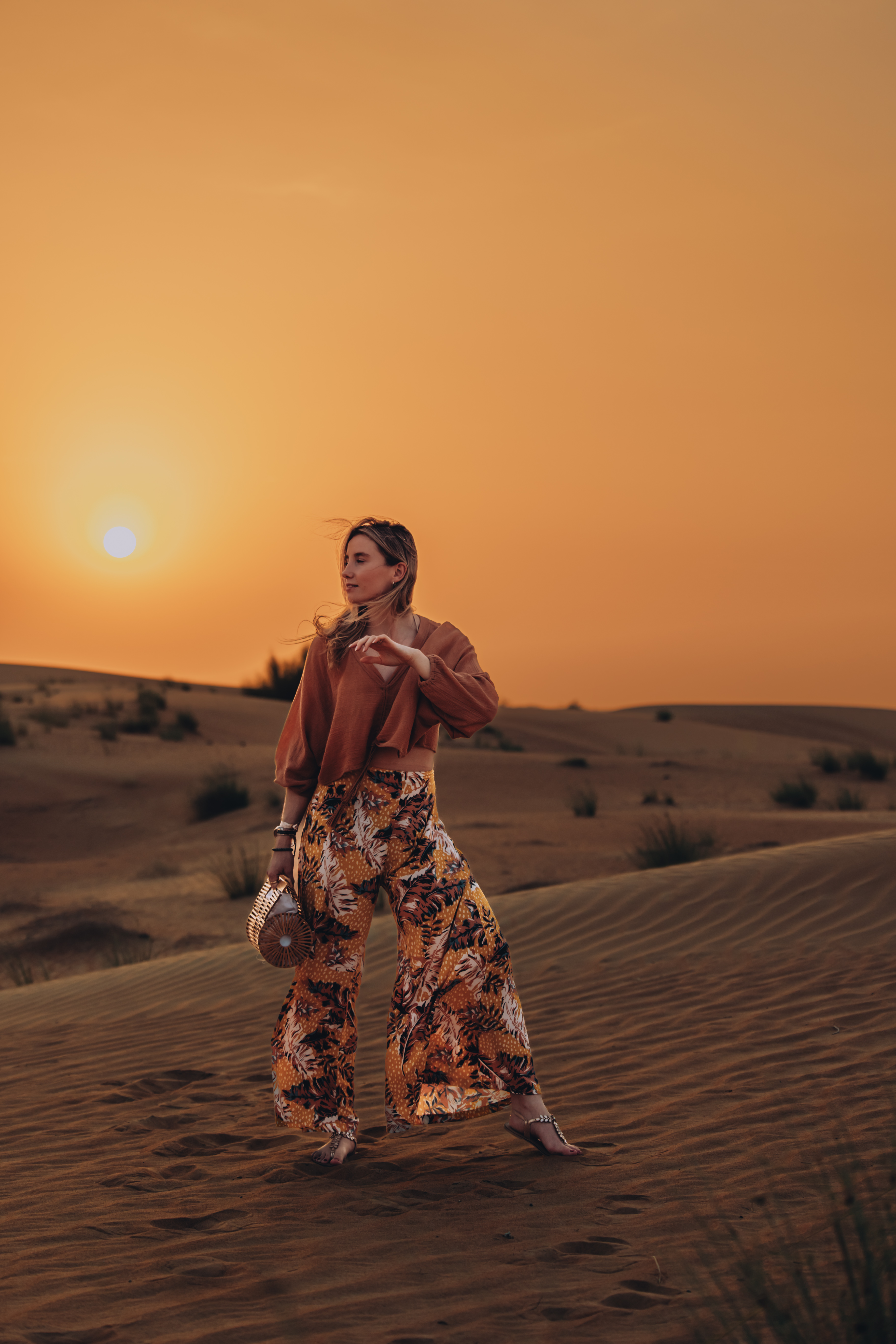 A woman in flowing printed trousers moving with the wind on desert dunes at sunset